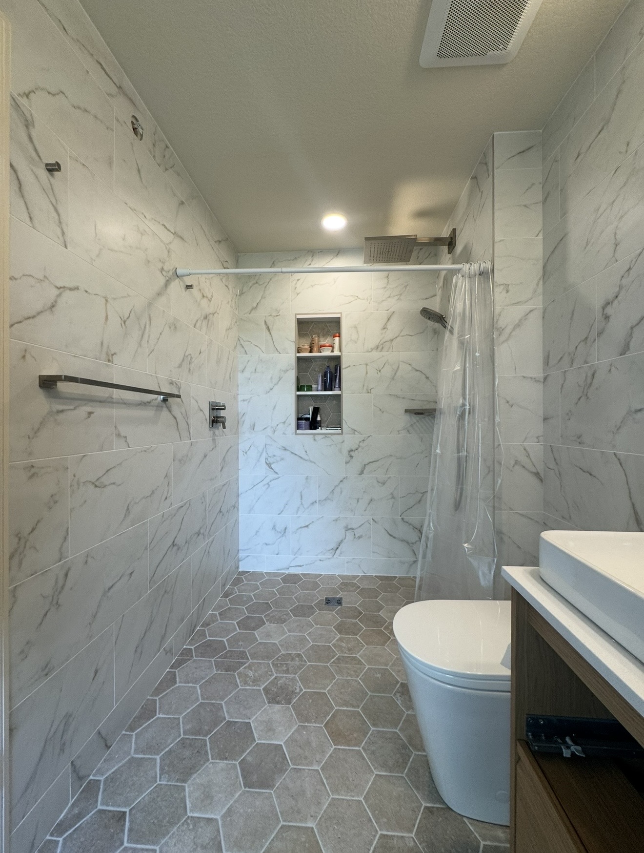 Curbless walk-in shower with marble-pattern wall tile, hexagonal floor tile, recessed niche, and brushed nickel fixtures