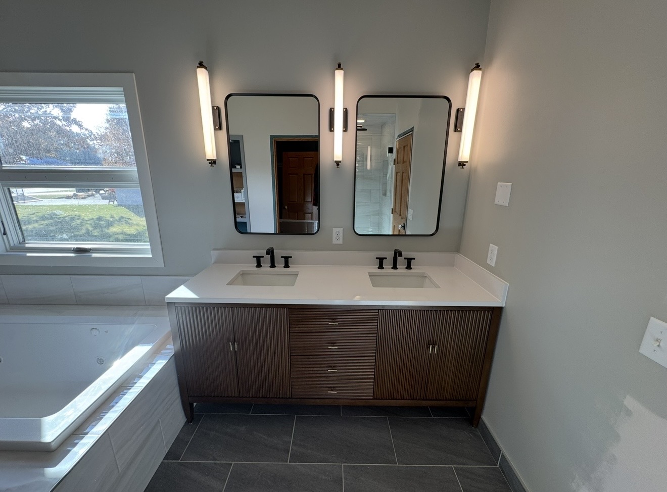 Double-sink bathroom with dark fluted wood vanity, white quartz countertop and backsplash, matte black faucets, vertical tube sconces, and dark floor tile