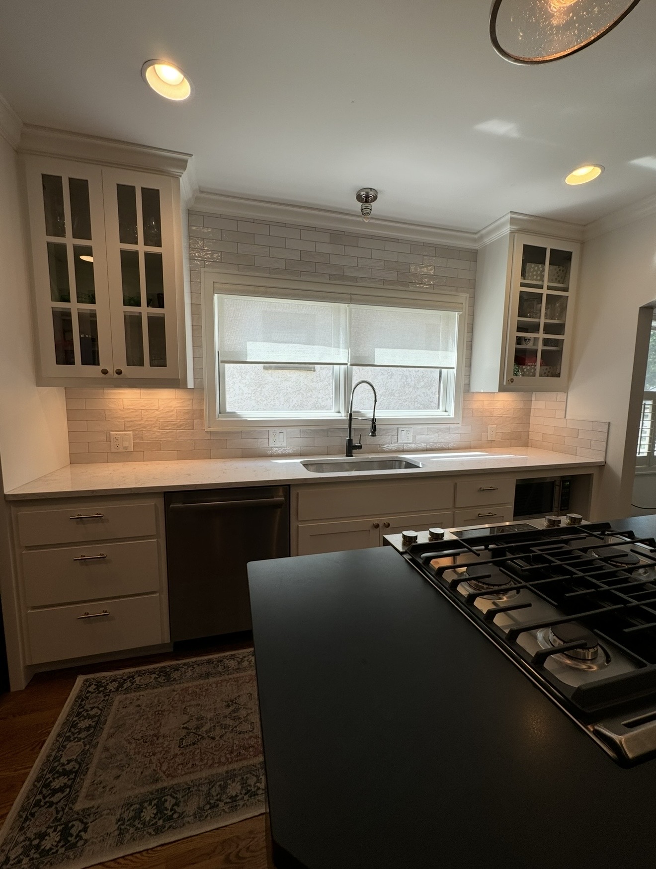 Bright kitchen with white cabinetry, glass-front upper cabinets, white subway tile backsplash, marble-style countertops, window over sink, and island with gas cooktop