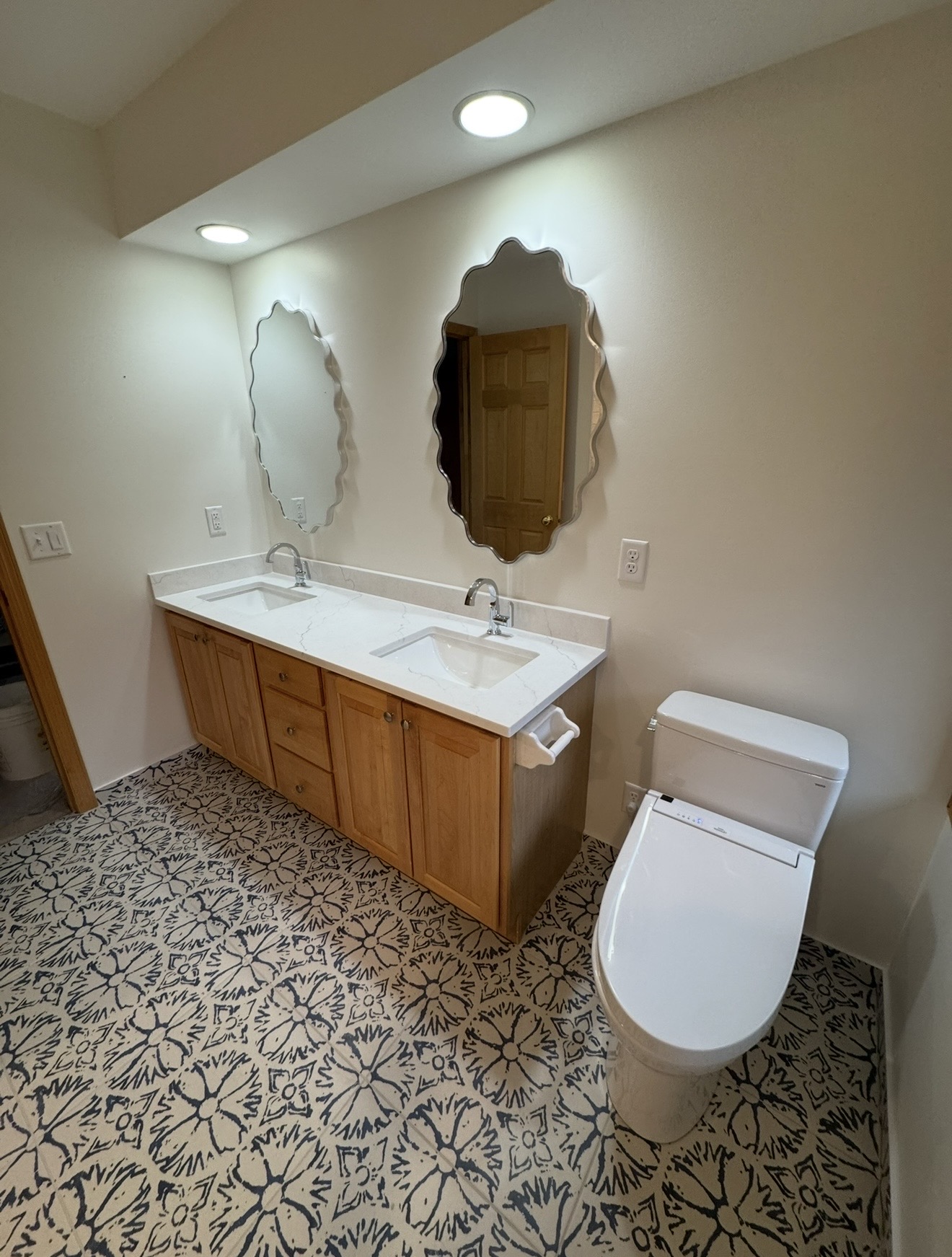 Remodeled bathroom with blue floral patterned floor tile, light wood double vanity, white quartz countertops, and scalloped mirrors