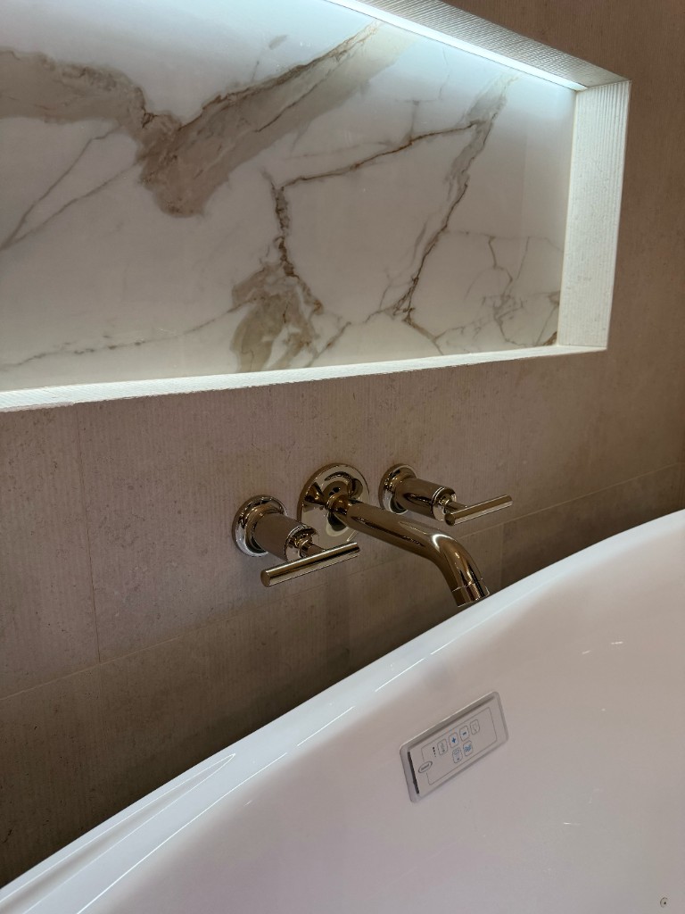 Wall-mounted tub filler with chrome fixtures, LED-lit niche with marble back, textured stone wall tile, and white tub rim with digital controls
