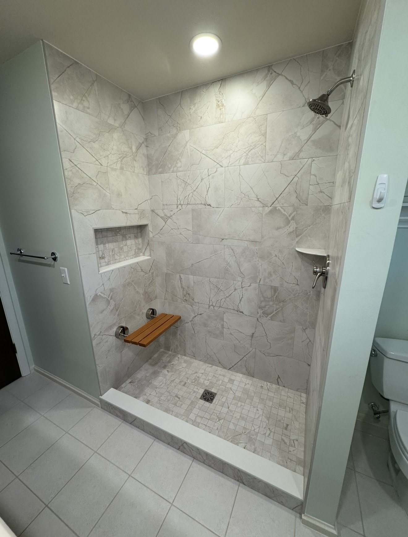Walk-in shower with marble-pattern wall and mosaic floor tile, recessed niche, fold-down wood bench, white curb, brushed metal fixtures, and pale mint green bathroom walls