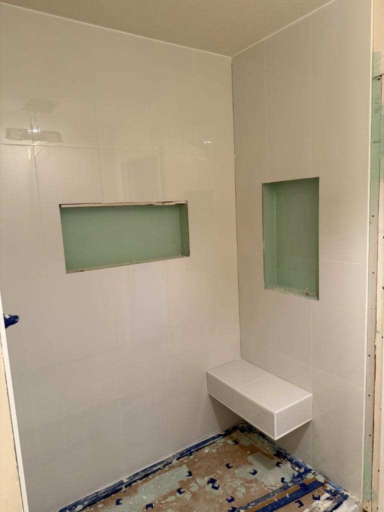 Walk-in shower under construction with glossy white large-format wall tile, horizontal and vertical recessed niches, floating corner bench, and waterproofing visible on floor