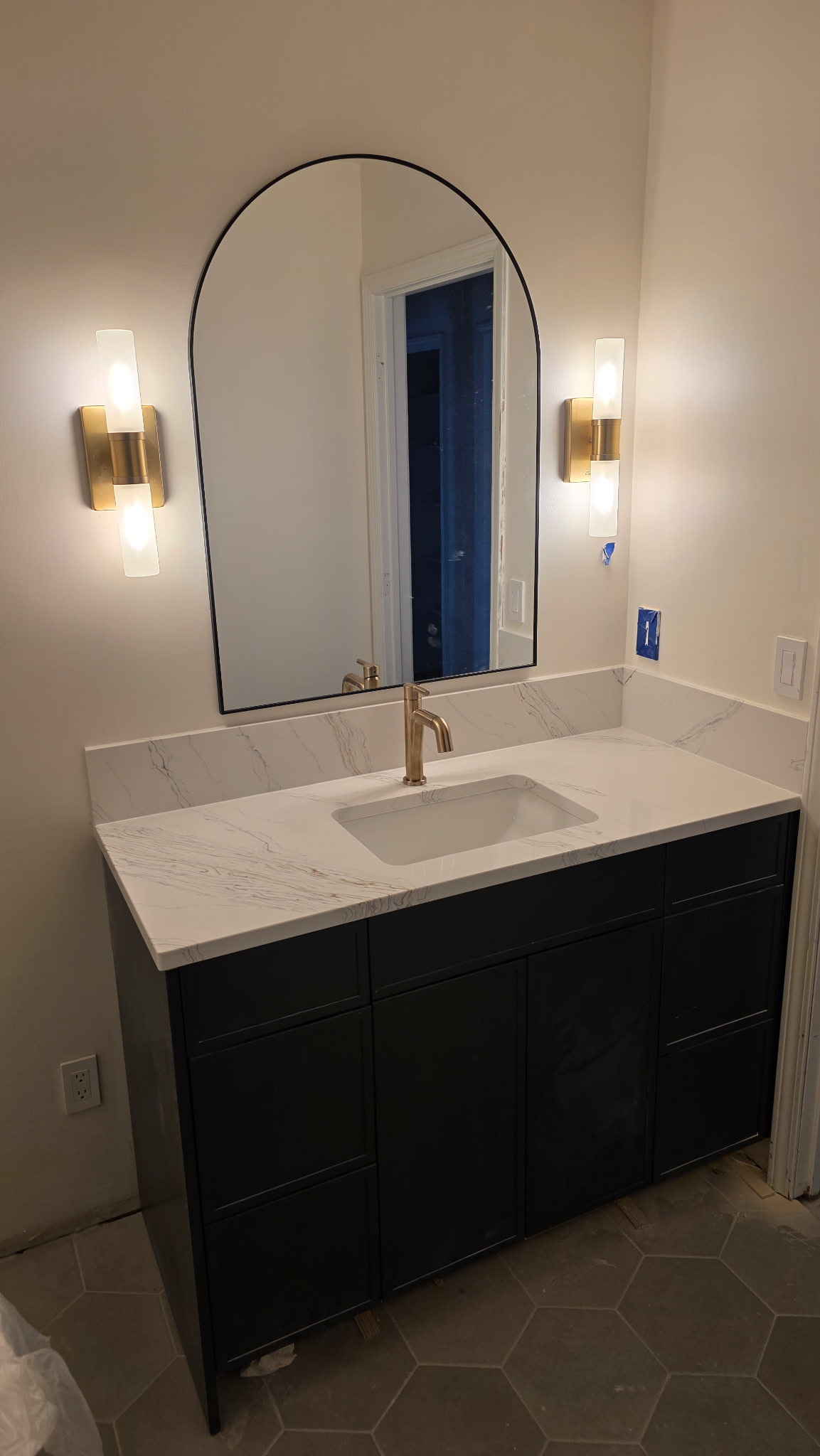 Bathroom remodel with dark vanity, marble-look countertop and backsplash, brushed gold faucet and sconces, arched mirror, and gray hex floor tile